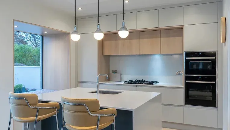 Modern minimalist kitchen with white island and integrated sink, pendant lighting, light wood cabinetry, built-in double oven, and large aluminium picture window - Senator Windows